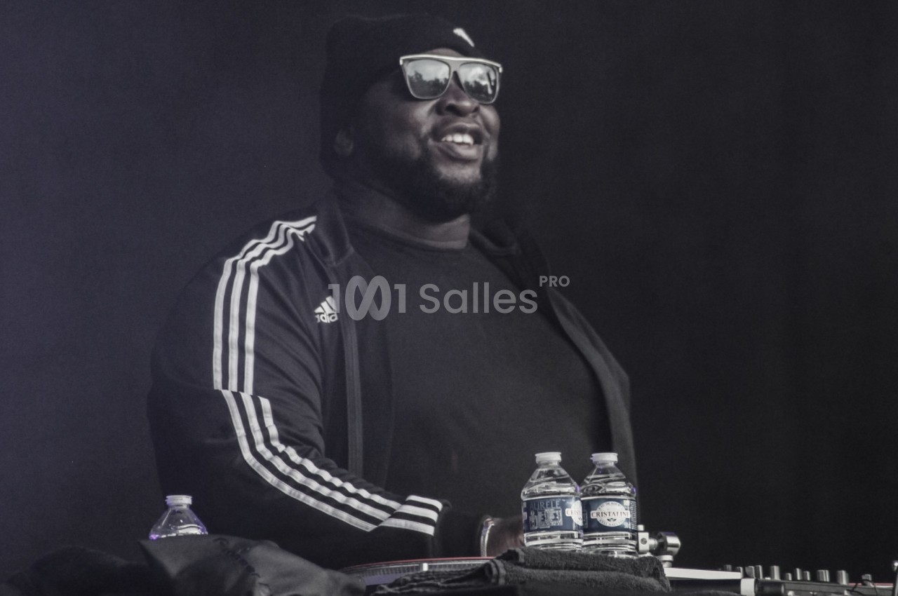 Un DJ portant des lunettes de soleil et un bonnet mixe sur scène avec des bouteilles d'eau posées devant lui.