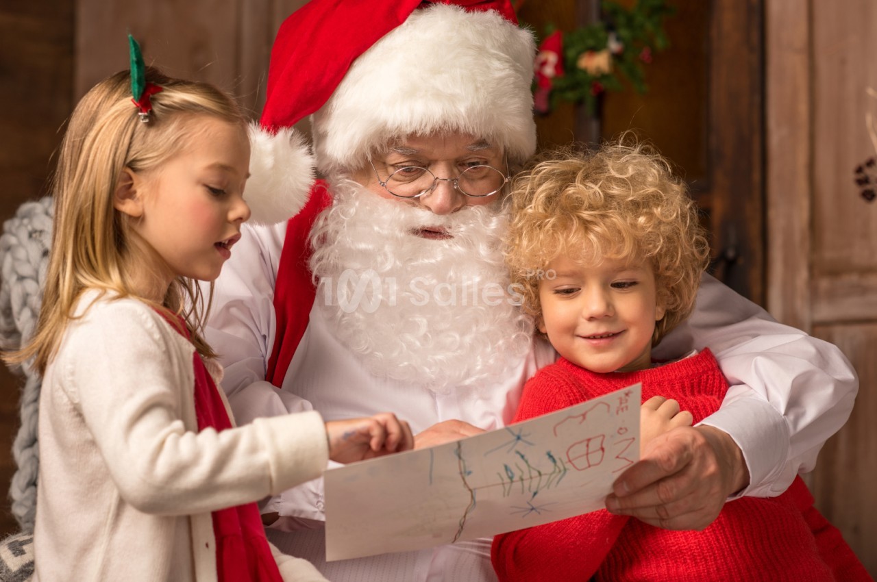 Un homme déguisé en Père Noël lit une lettre avec deux enfants souriants dans un décor chaleureux.