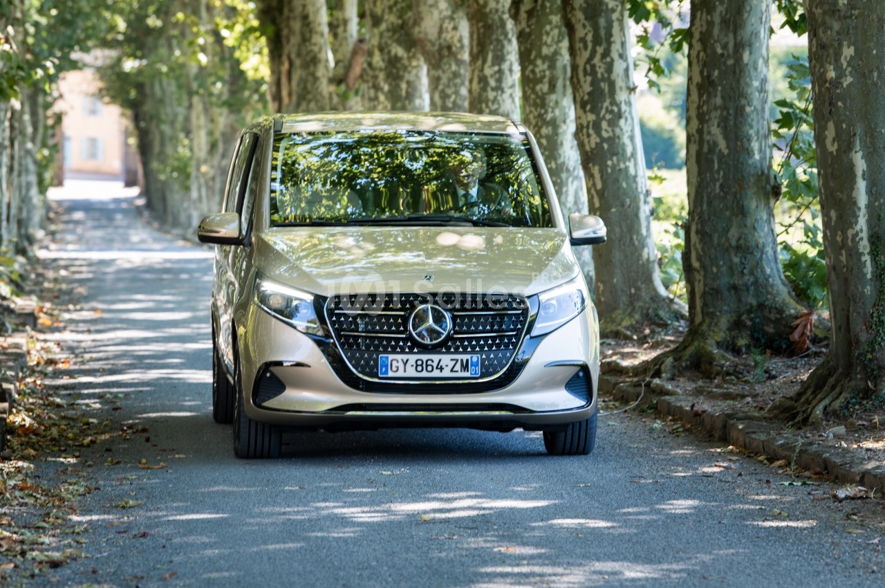 Monospace argenté Mercedes-Benz roulant sur une route bordée d'arbres, sous une lumière tamisée.
