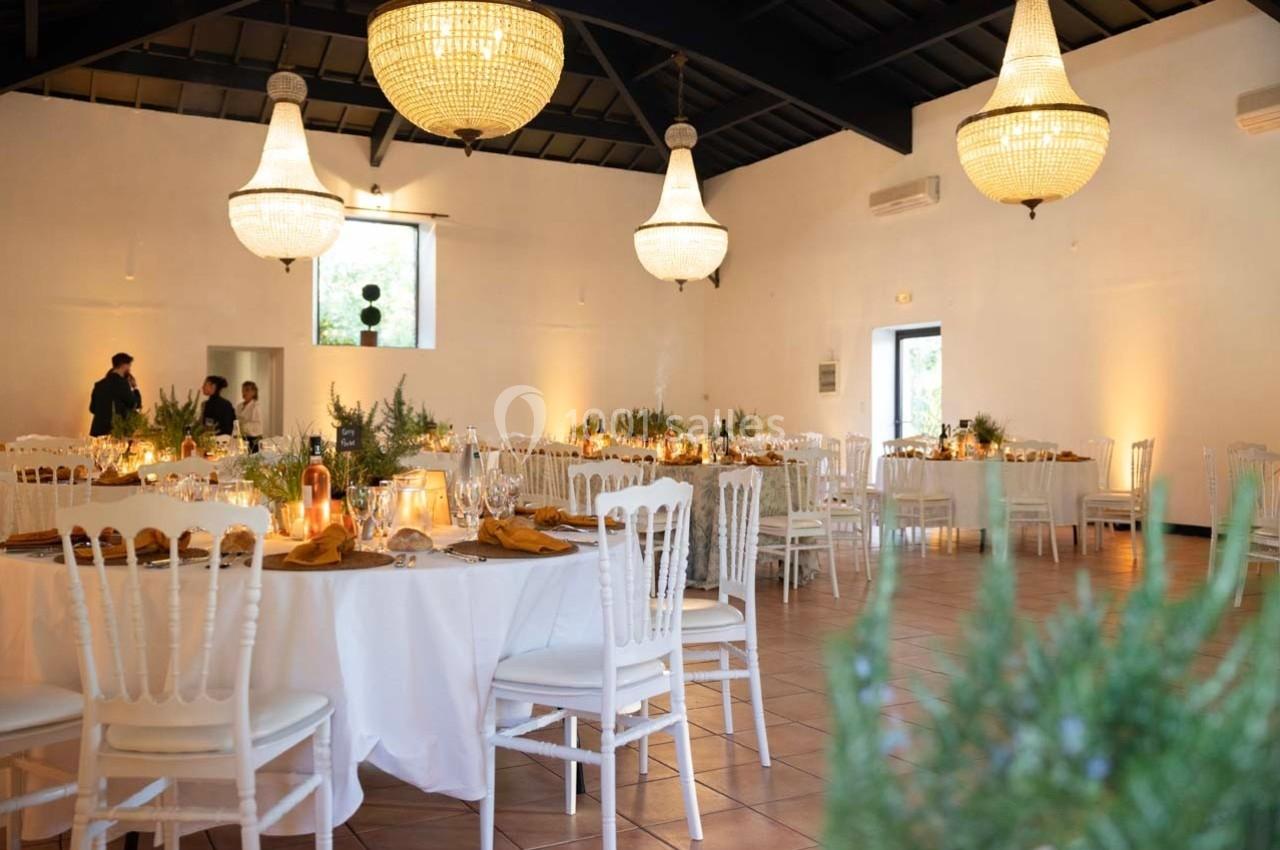 Salle de réception élégante avec tables rondes décorées, chaises blanches et grands lustres suspendus.