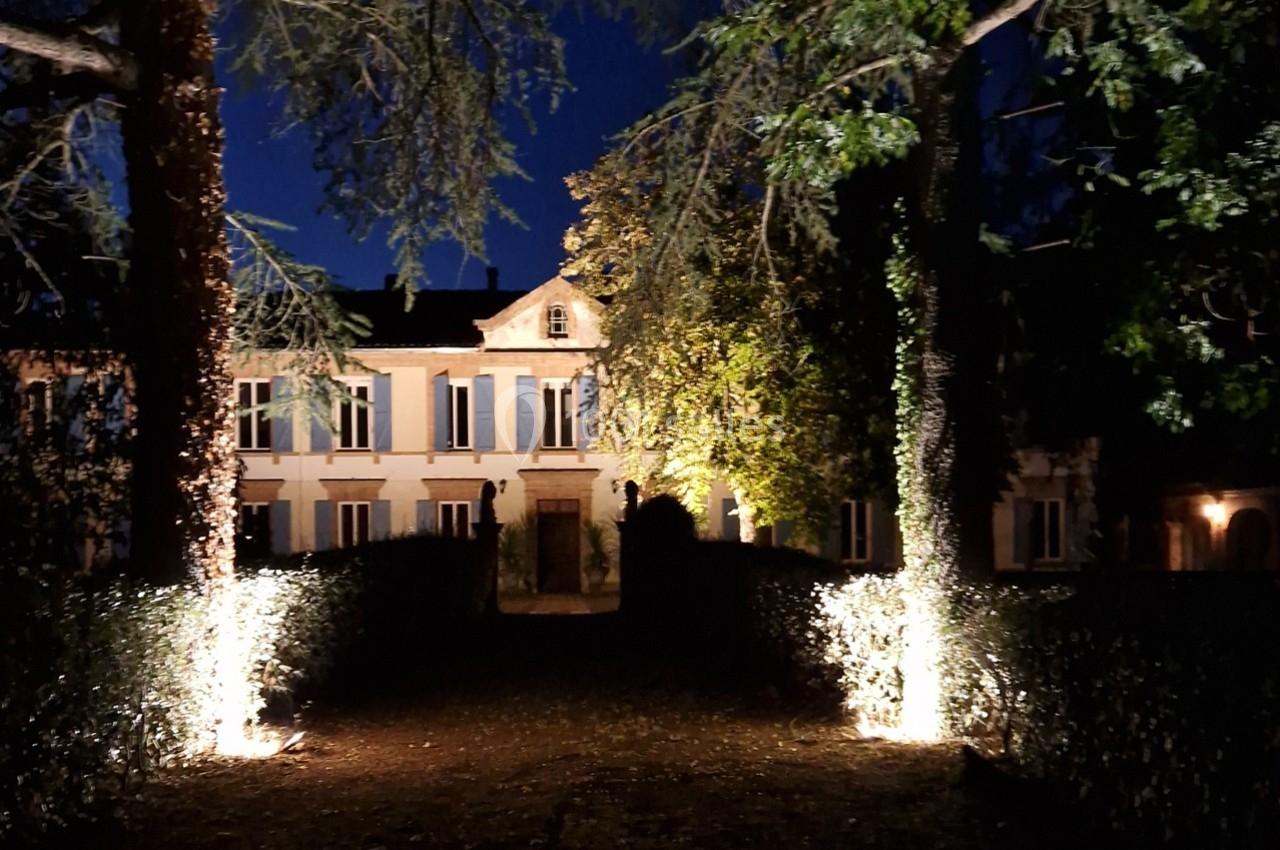 Façade d'une grande maison éclairée la nuit, entourée d'arbres et de haies dans un jardin sombre.