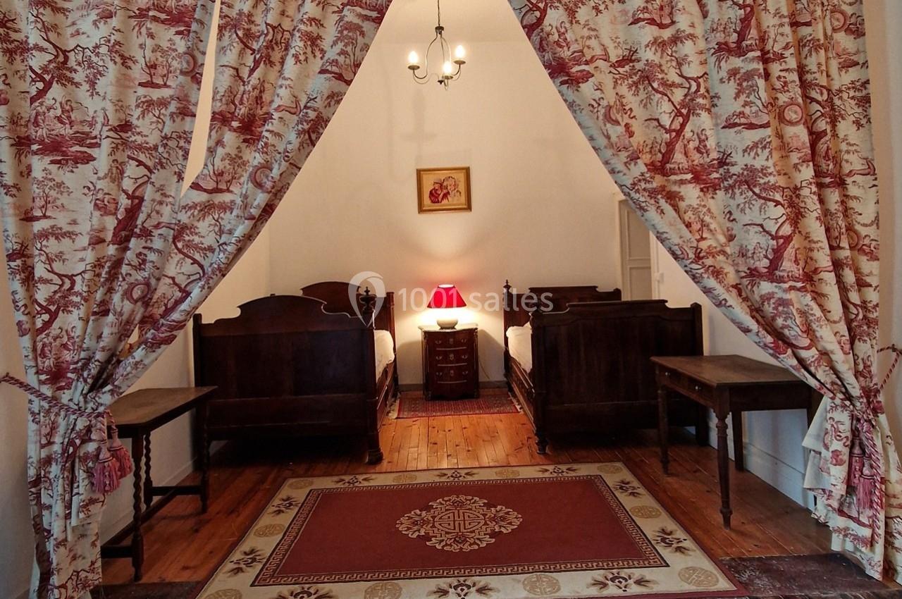 Deux lits en bois ancien dans une chambre avec rideaux à motifs, tapis rouge et lampe de chevet allumée.