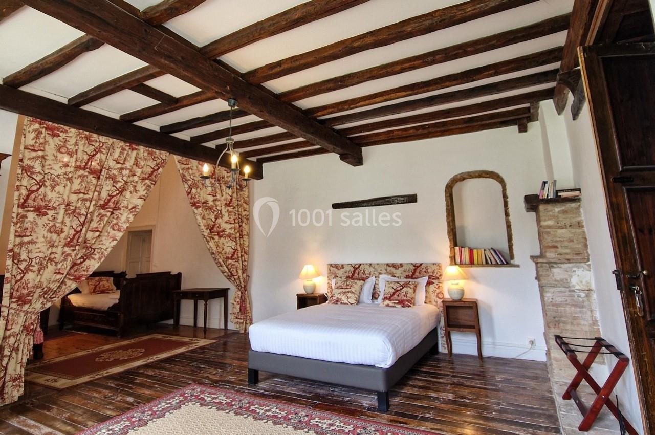 Chambre avec poutres apparentes, lit double, rideaux en toile de Jouy, mobilier en bois et tapis sur parquet.