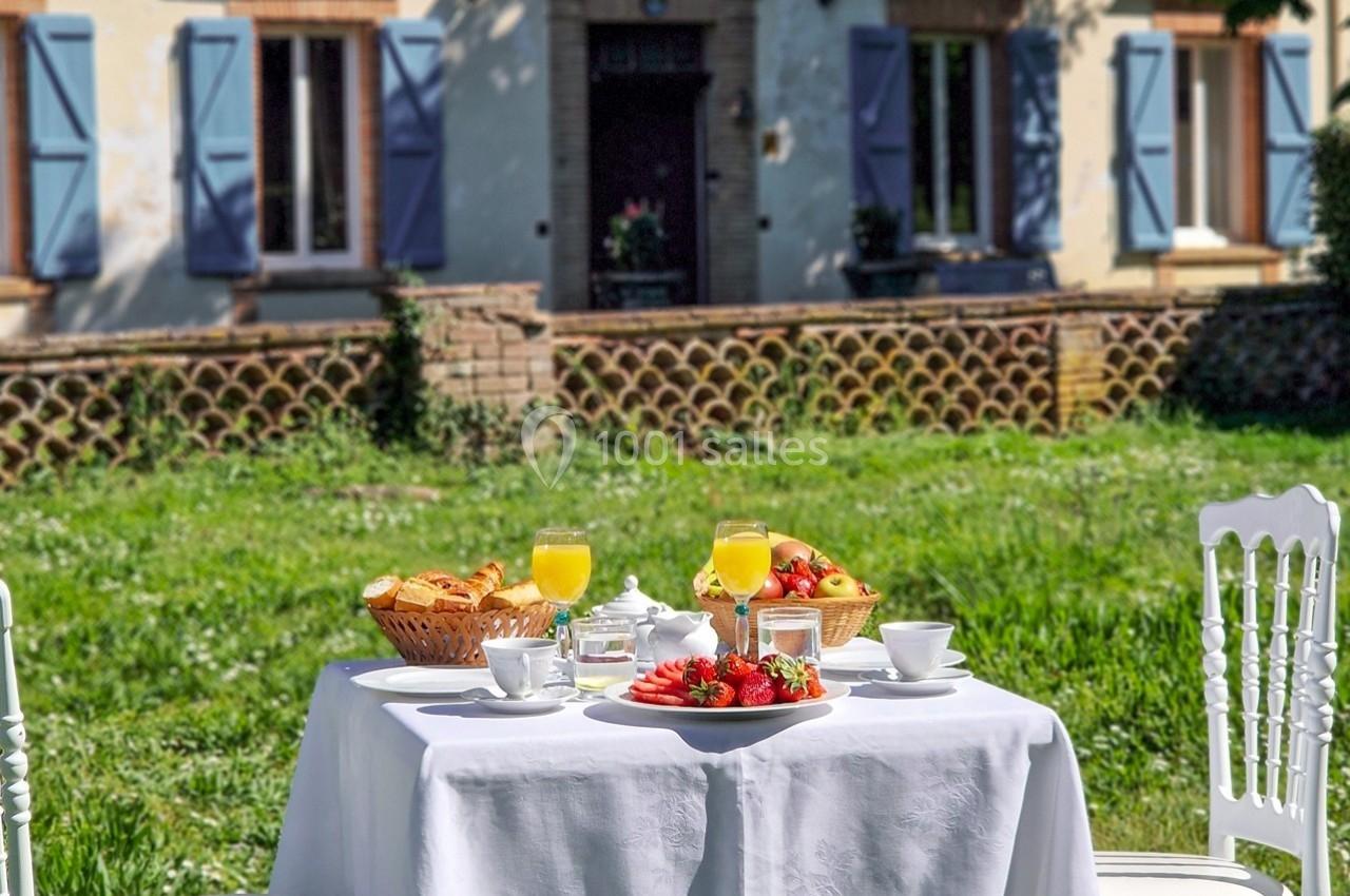 Table dressée en extérieur avec petit-déjeuner composé de fruits, viennoiseries et jus d'orange, devant une maison.