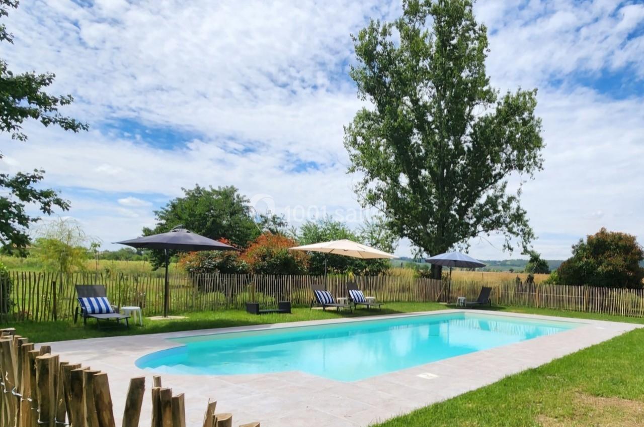 Piscine extérieure entourée de chaises longues, parasols, pelouse et arbres sous un ciel partiellement nuageux.