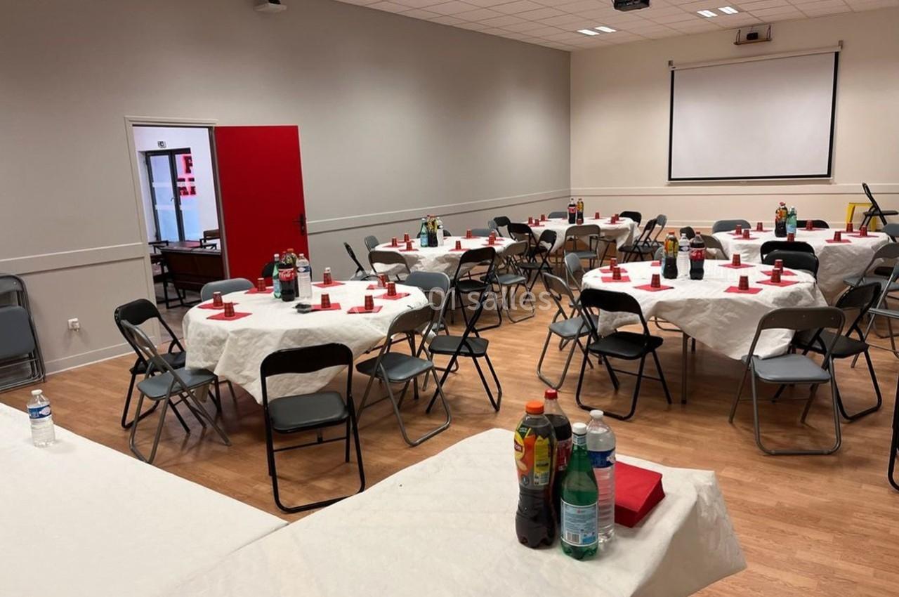 Salle aménagée avec des tables rondes dressées, nappes blanches, chaises pliantes et boissons disposées.