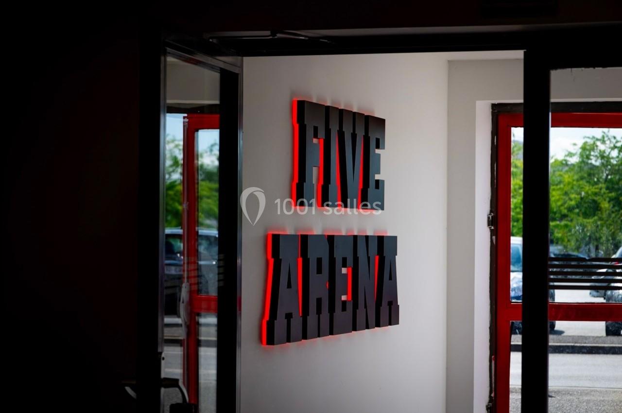 Enseigne lumineuse rouge et noire indiquant ’Five Arena’ sur un mur blanc près d'une porte ouverte.