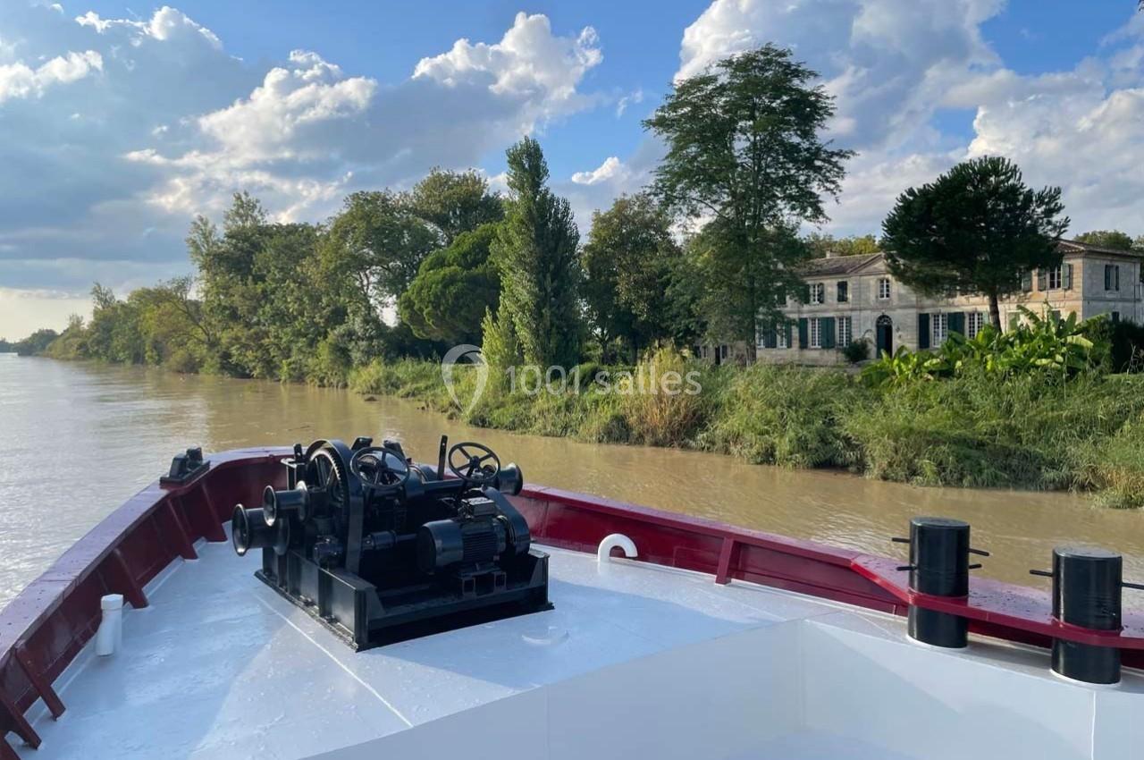 Vue depuis l'avant d'un bateau naviguant sur une rivière bordée d'arbres et d'une maison en pierre.