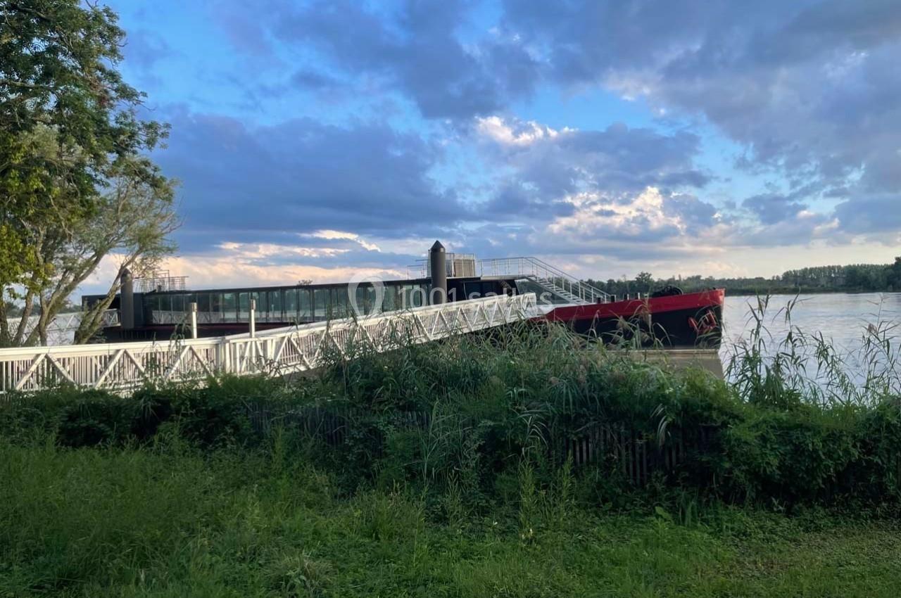 Passerelle menant à un bateau amarré sur une rivière, entourée de végétation et sous un ciel partiellement nuageux.