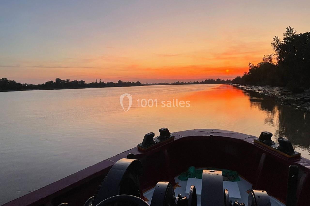 Vue d'un fleuve au coucher du soleil depuis un bateau, avec des arbres bordant les rives et un ciel orangé.