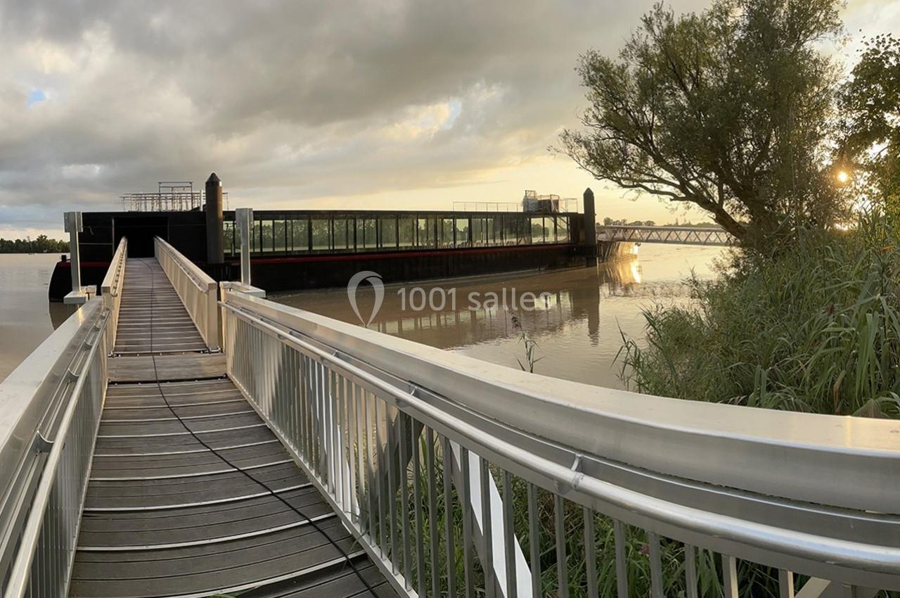 Passerelle en bois et métal menant à une péniche amarrée sur une rivière, avec un pont visible à l'arrière-plan.