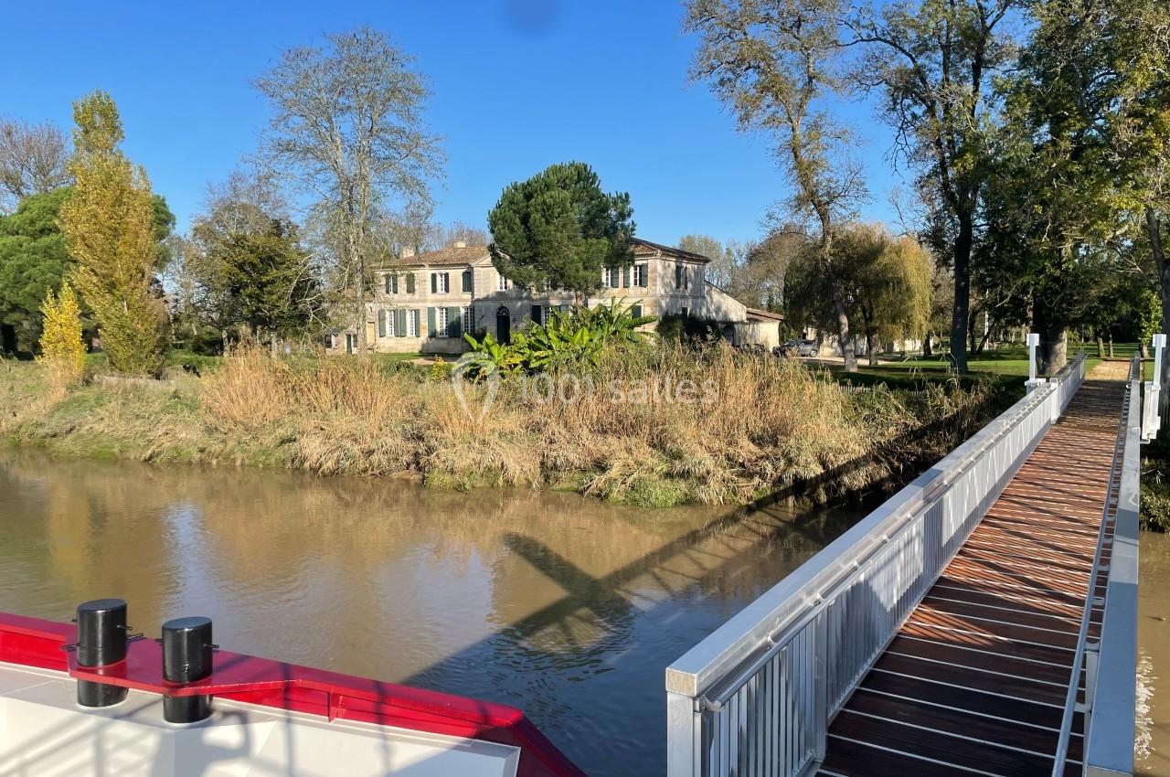 Vue d'une maison en pierre entourée d'arbres, bordant un canal avec une passerelle en bois au premier plan.