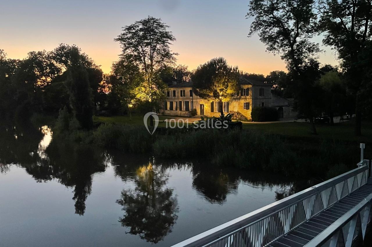 Vue d'un bâtiment éclairé au bord d'une rivière au crépuscule, entouré d'arbres et reflété dans l'eau calme.
