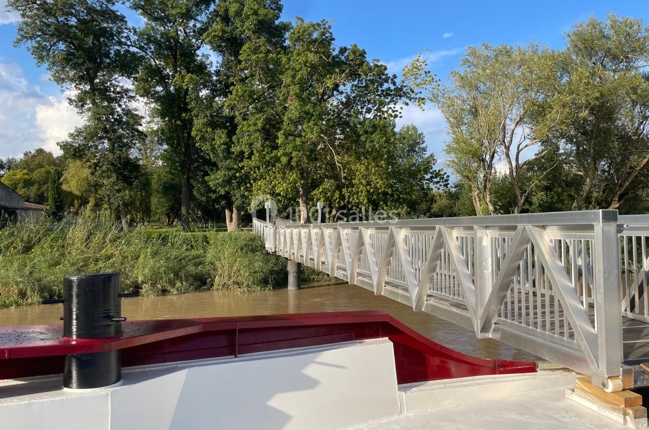 Passerelle en bois et métal traversant une rivière, entourée de végétation, vue depuis un bateau.