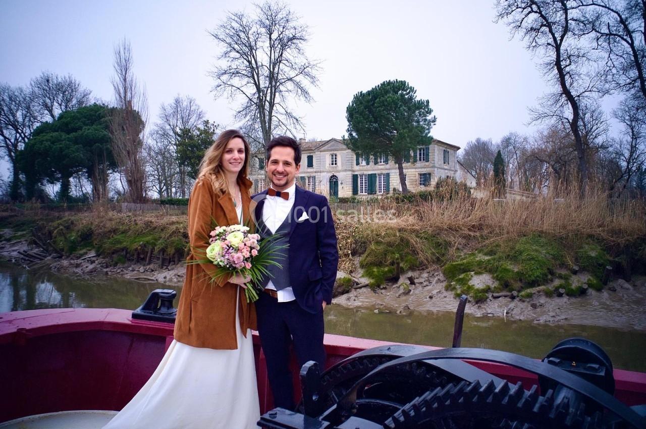 Un couple en tenue de mariage pose sur un bateau devant une maison en pierre entourée d'arbres en hiver.