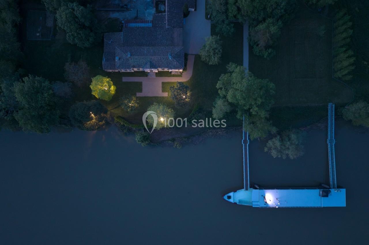 Vue aérienne nocturne d'une maison éclairée près d'une rivière avec un ponton et un bateau amarré.