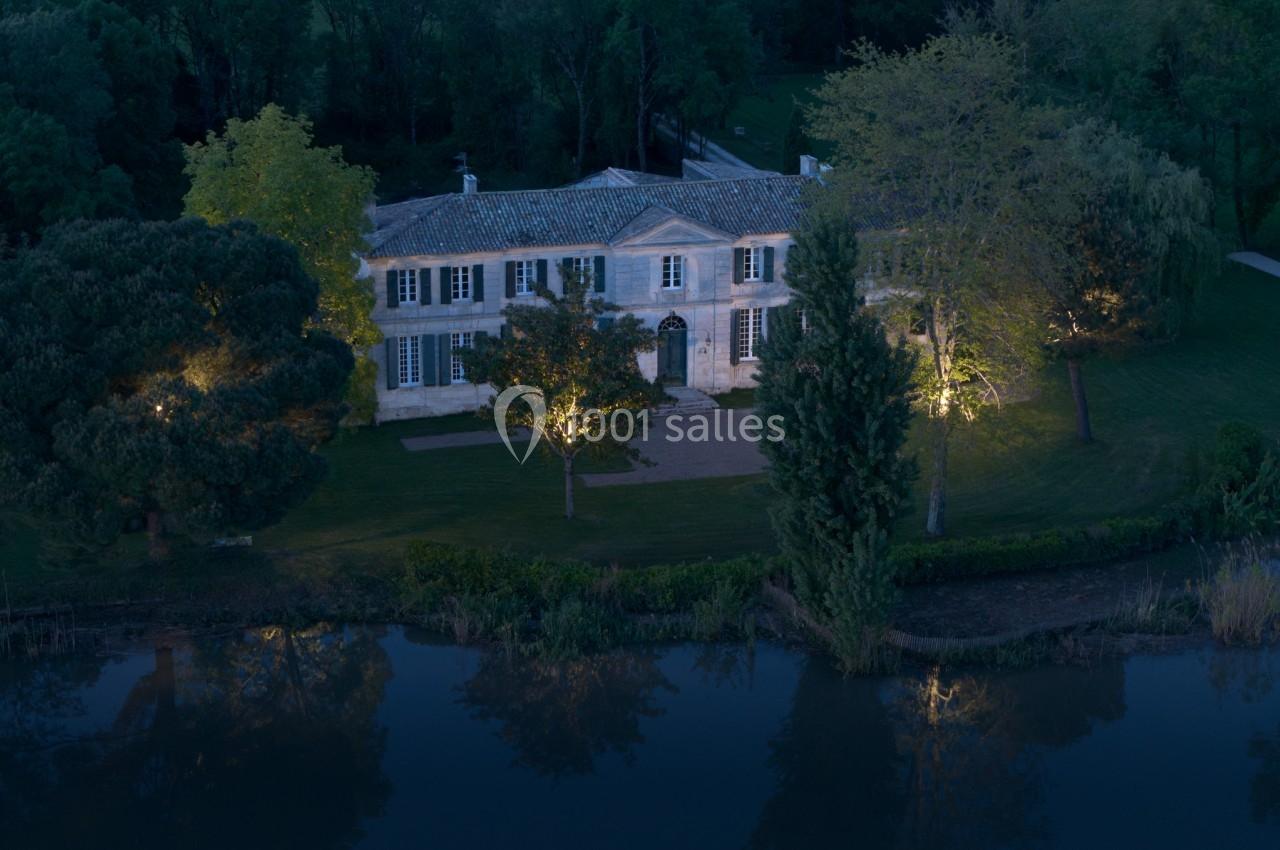 Vue nocturne d'une grande maison en pierre entourée d'arbres, avec un jardin éclairé et un étang au premier plan.
