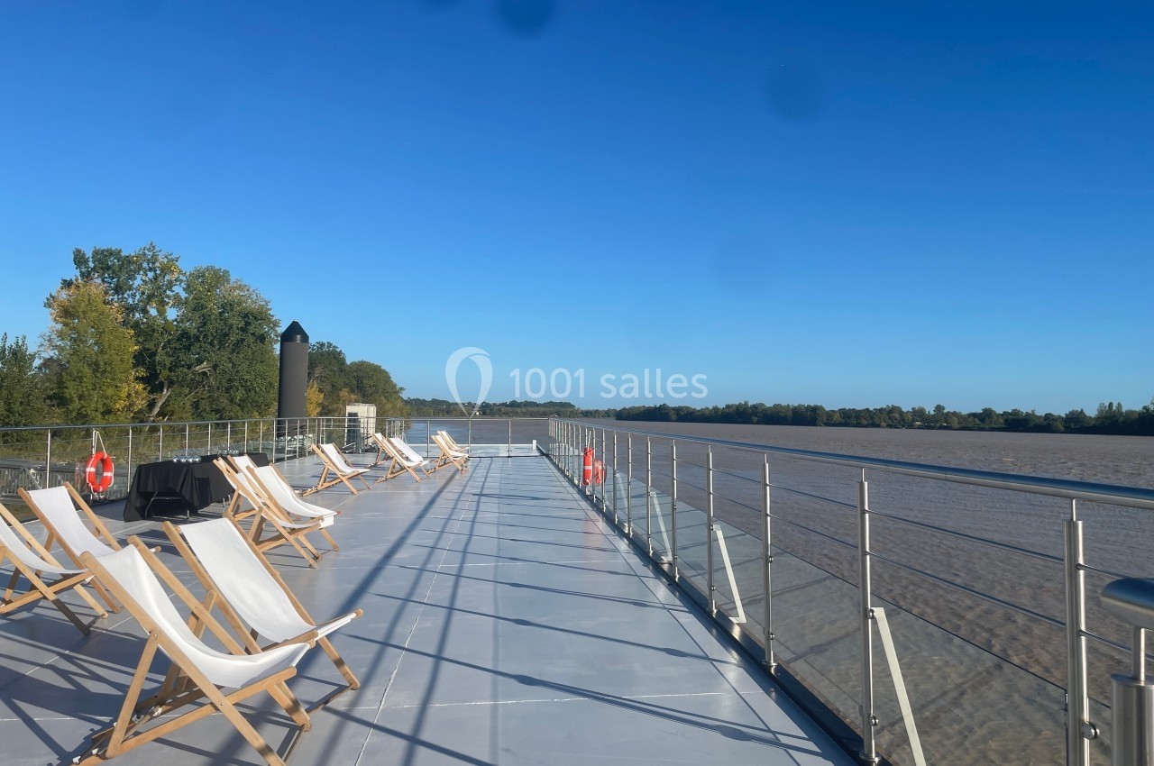Terrasse d'un bateau avec des transats alignés, vue sur un fleuve et des arbres sous un ciel bleu dégagé.