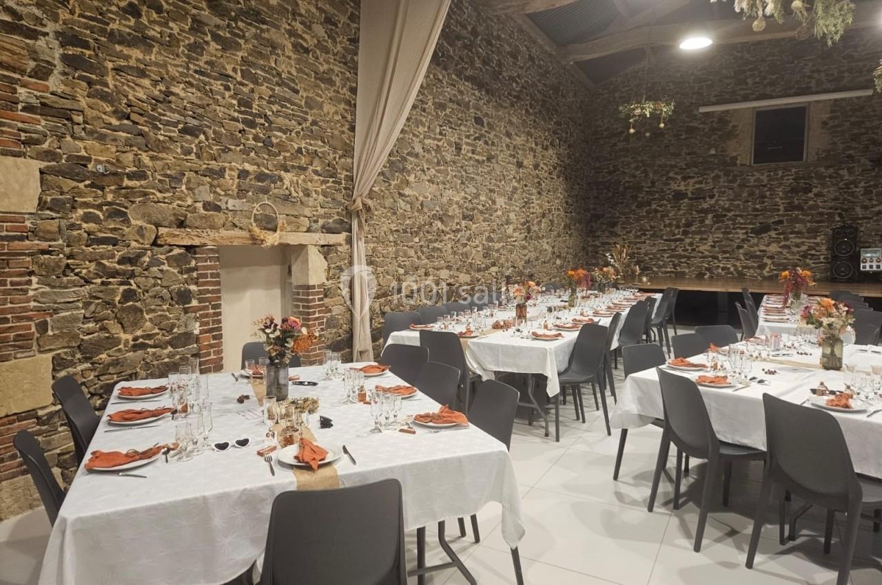 Salle en pierre aménagée pour un repas, avec tables dressées, nappes blanches, vaisselle et décorations florales.
