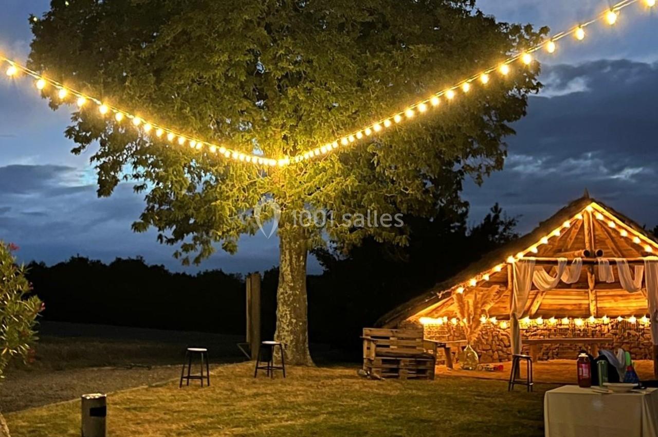 Guirlandes lumineuses éclairant un arbre et une grange en bois dans un cadre extérieur au crépuscule.