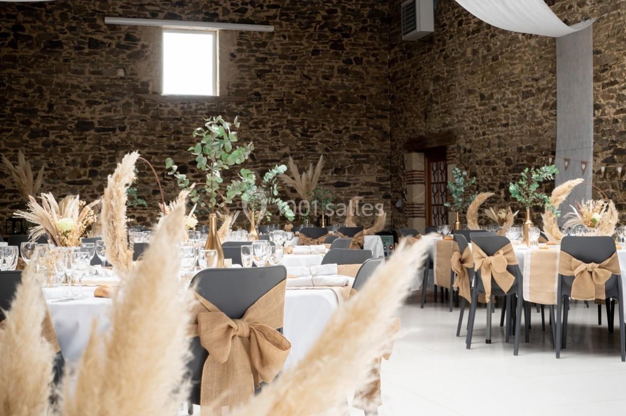 Salle de réception décorée avec des tables dressées, nappes blanches, chaises ornées de nœuds en toile de jute et bouquets…