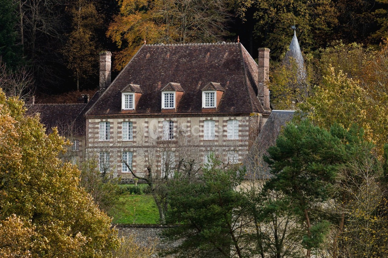 Manoir en pierre entouré d'arbres et de verdure, avec un toit en tuiles et des fenêtres à volets blancs.