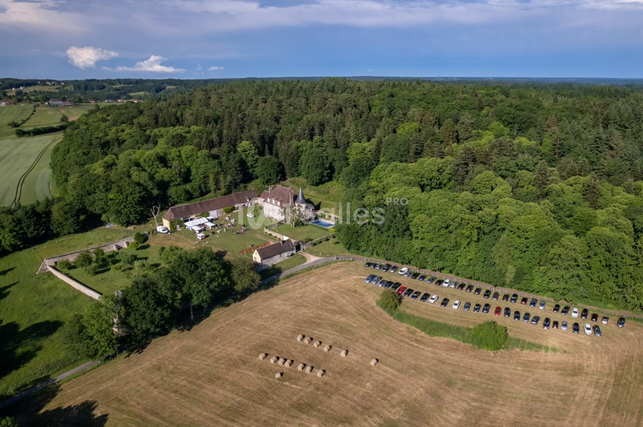 Vue aérienne d'une ferme entourée de forêt, avec des champs, des bottes de foin et un parking rempli de voitures.