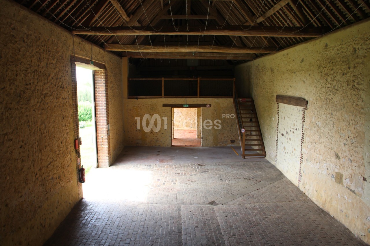 Intérieur d'une grange en pierre avec un sol en briques, une grande porte ouverte et un escalier en bois sur le côté.
