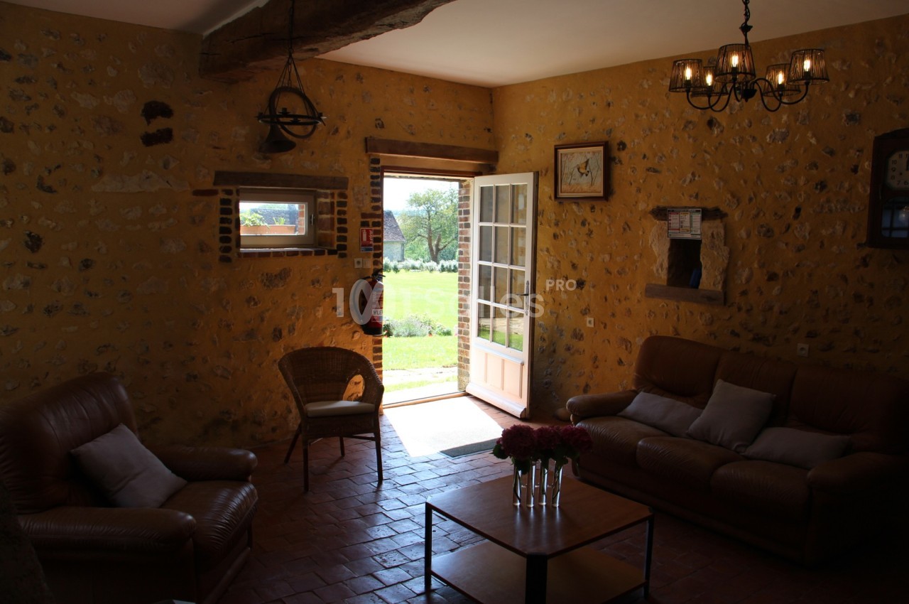 Salon rustique avec murs en pierre, canapés en cuir, table basse et porte ouverte donnant sur un jardin ensoleillé.