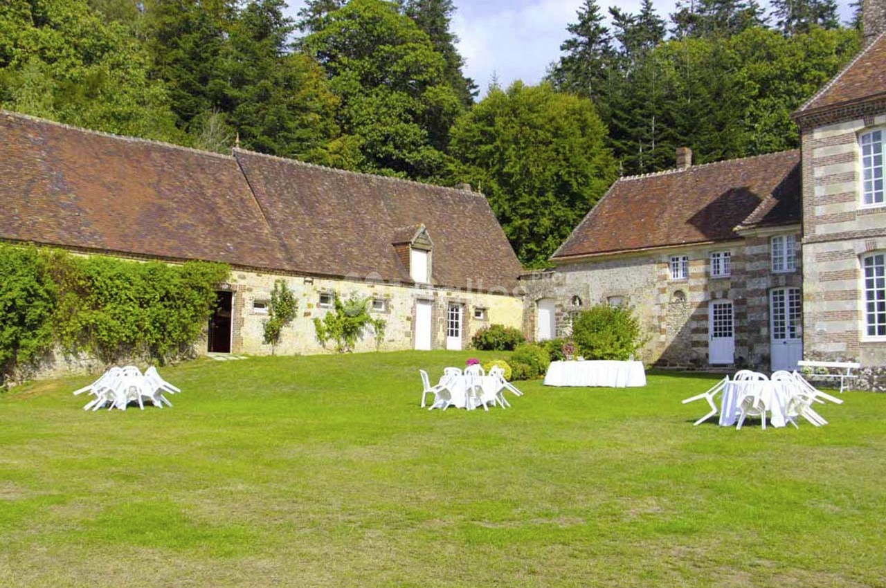 Cour verdoyante avec bâtiments en pierre, chaises et tables blanches disposées pour un événement en extérieur.