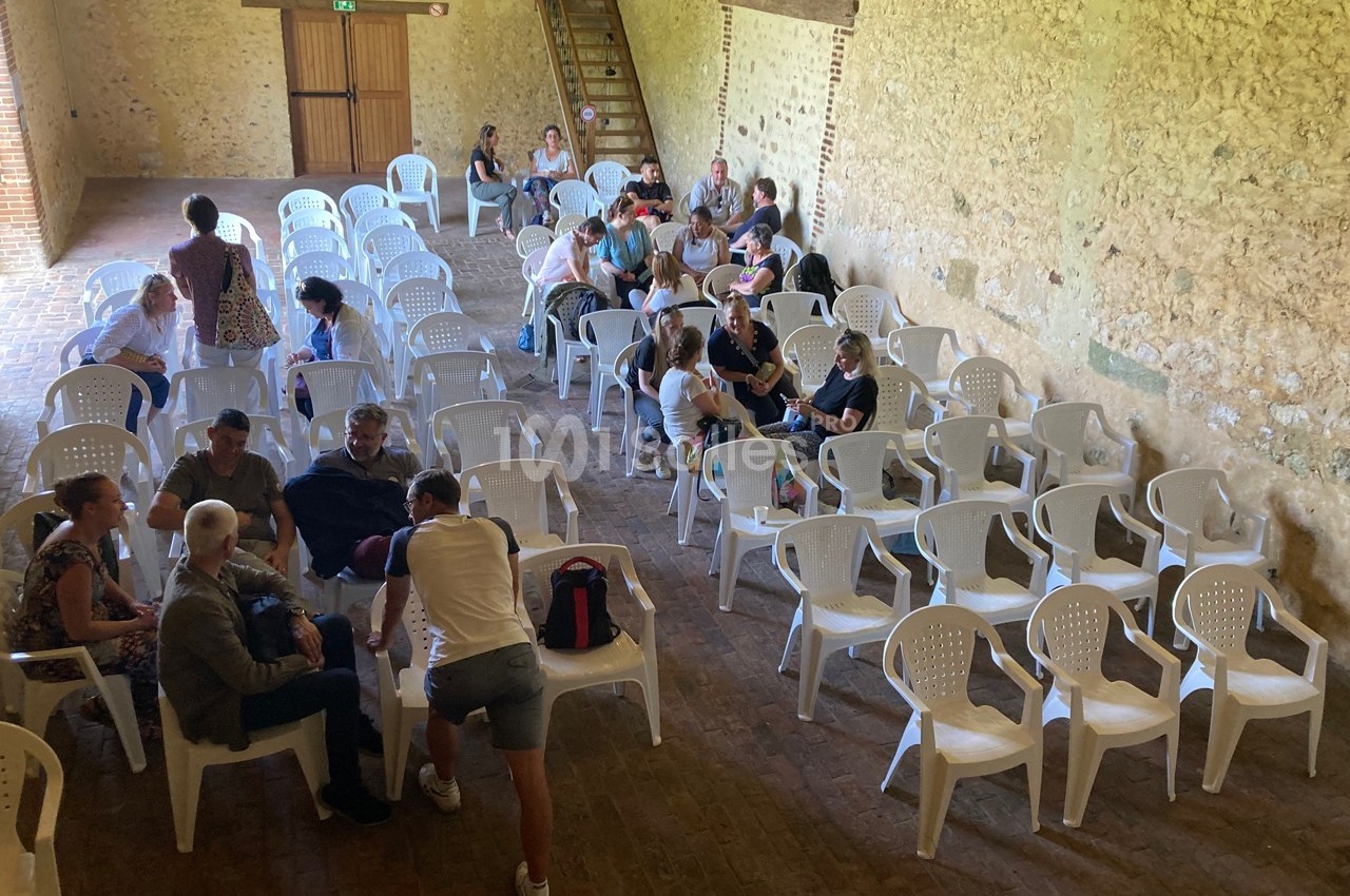 Des personnes assises et discutant dans une salle avec des murs en pierre et des chaises en plastique blanc.