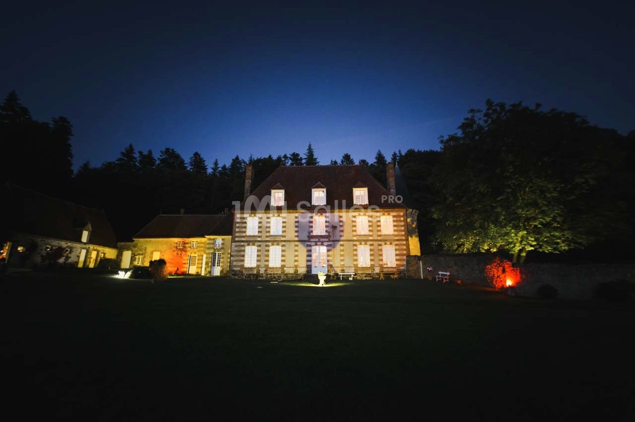 Façade éclairée d'un grand bâtiment ancien entouré d'arbres, photographiée de nuit avec un éclairage coloré.