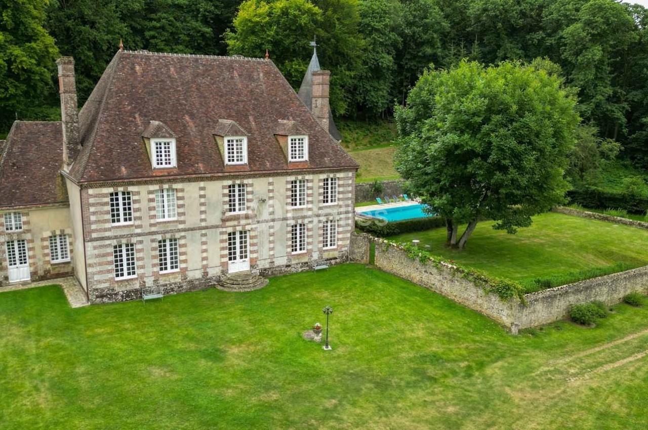 Manoir en pierre avec toit en ardoise, grand jardin verdoyant, piscine et forêt en arrière-plan.