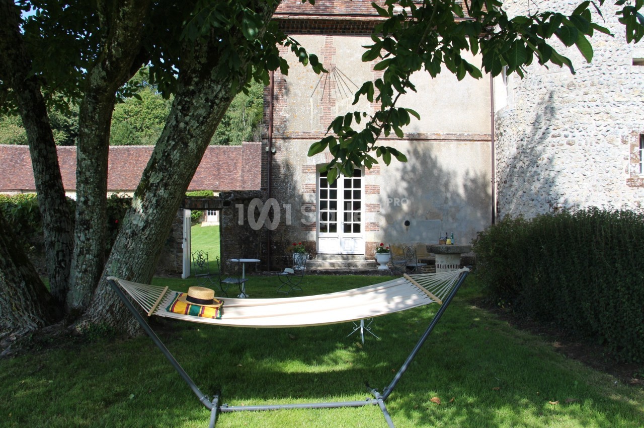 Hamac beige installé sous un arbre dans un jardin verdoyant, près d'une maison en pierre avec des volets blancs.