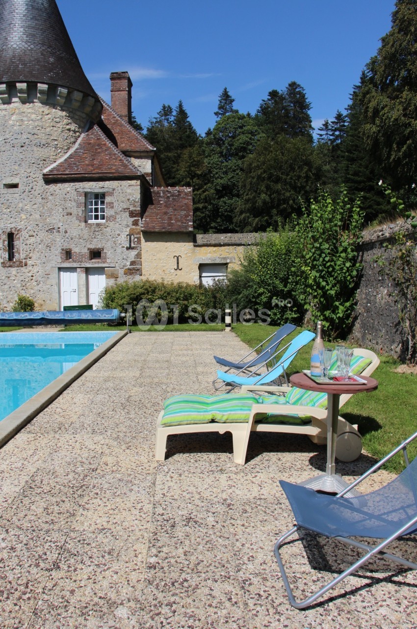 Chaises longues et table près d'une piscine, avec un bâtiment en pierre et des arbres en arrière-plan.