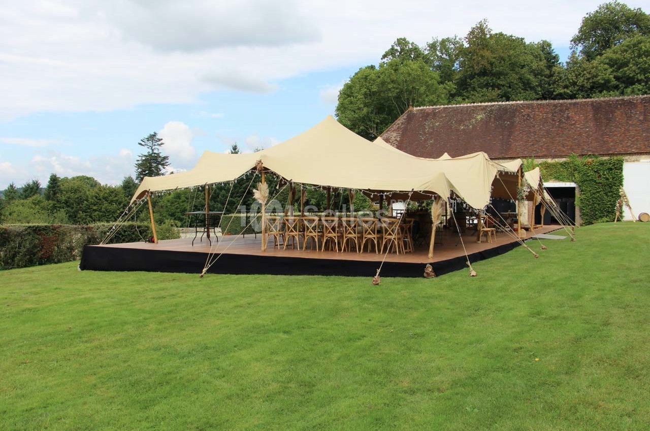 Tente beige montée sur une pelouse, équipée de tables et chaises en bois, entourée de verdure et d'un bâtiment en arrière…