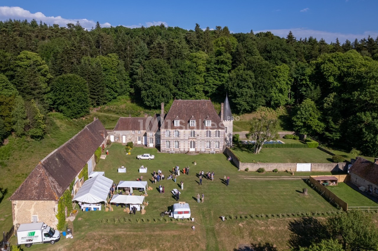 Vue aérienne d'un domaine avec bâtiments anciens, tentes blanches et personnes réunies dans un grand espace vert.