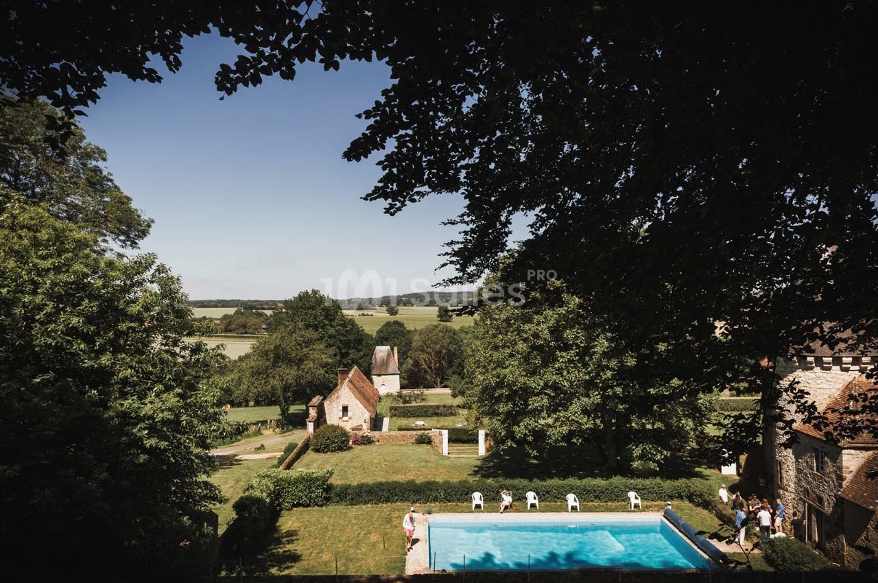 Vue d'un jardin avec piscine, entouré de bâtiments en pierre et de verdure, donnant sur une campagne vallonnée.