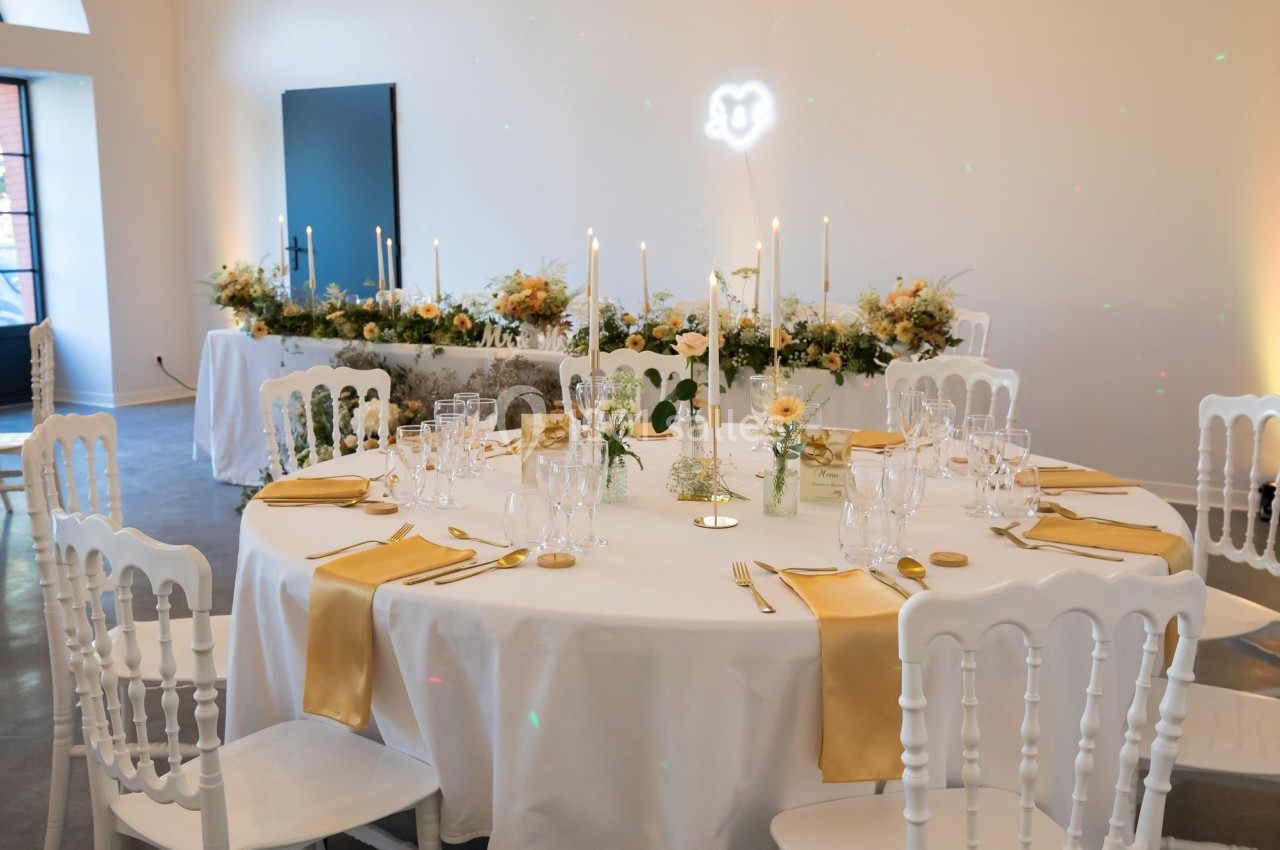 Table ronde décorée avec des nappes blanches, des serviettes dorées, des chandeliers et des fleurs, dans une salle lumineuse.
