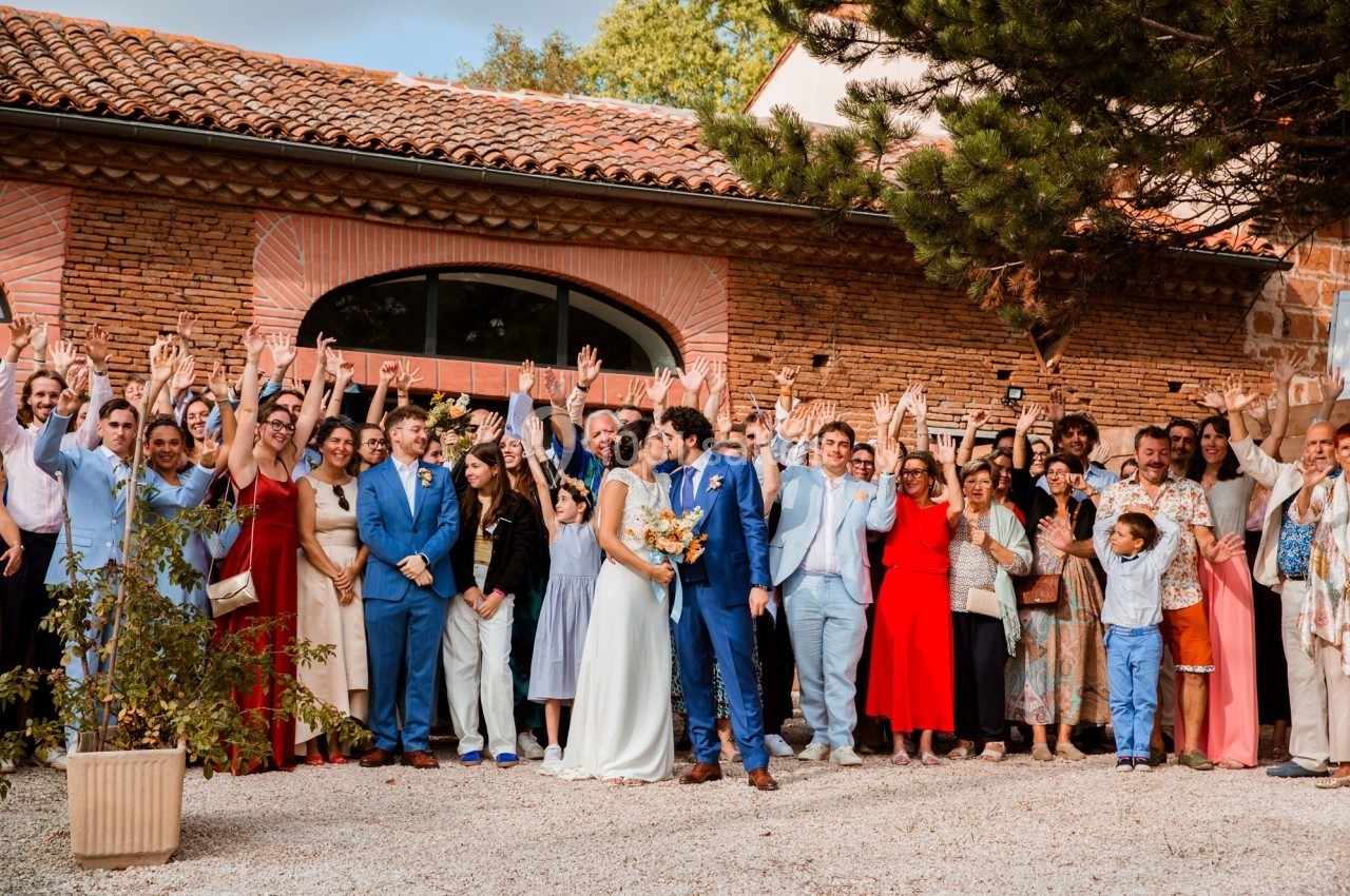 Un groupe de personnes pose joyeusement devant un bâtiment en briques, avec un couple au centre en tenue de mariage.