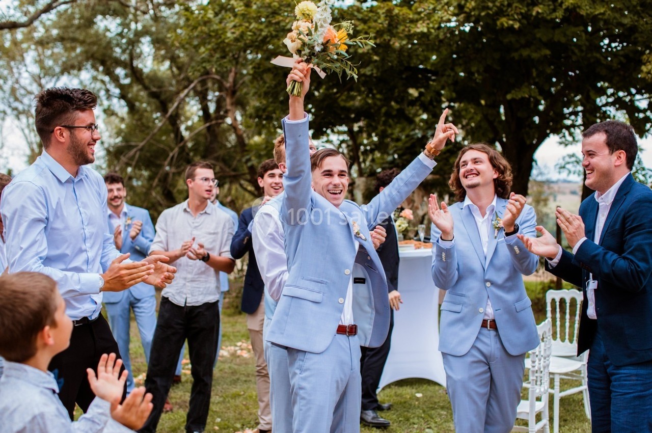 Un homme en costume clair lève un bouquet de fleurs, entouré de personnes applaudissant dans un cadre extérieur verdoyant.