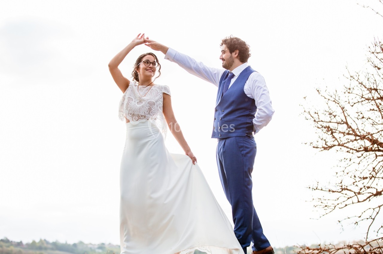 Un couple en tenue de mariage danse en extérieur, sous un ciel clair avec un arbre dénudé en arrière-plan.
