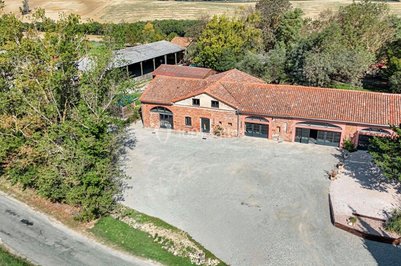 Bâtiment en briques rouges avec toit en tuiles, entouré d'arbres et de champs, vu depuis les airs.