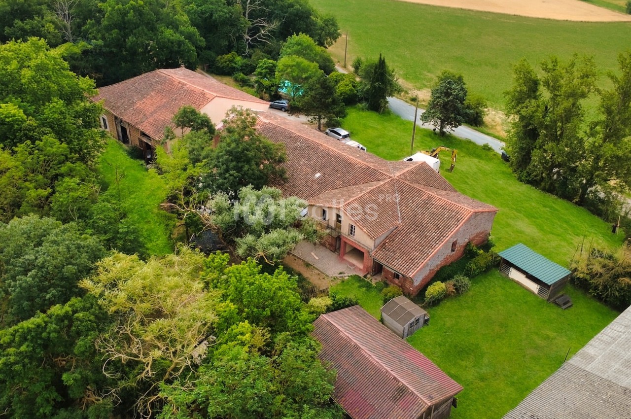 Vue aérienne d'une grande maison en briques avec toit en tuiles, entourée de verdure et de bâtiments annexes.