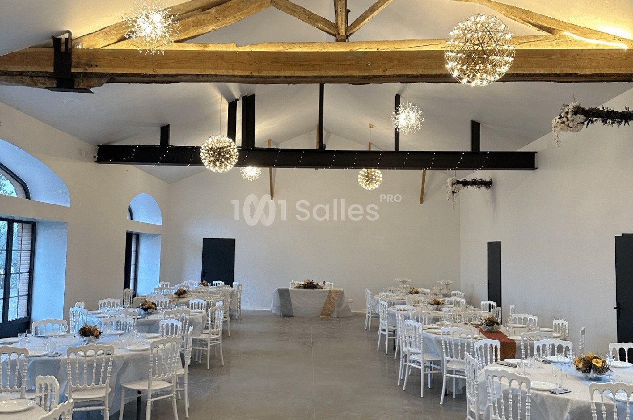 Salle de réception lumineuse avec poutres apparentes, tables rondes dressées et décorations florales.