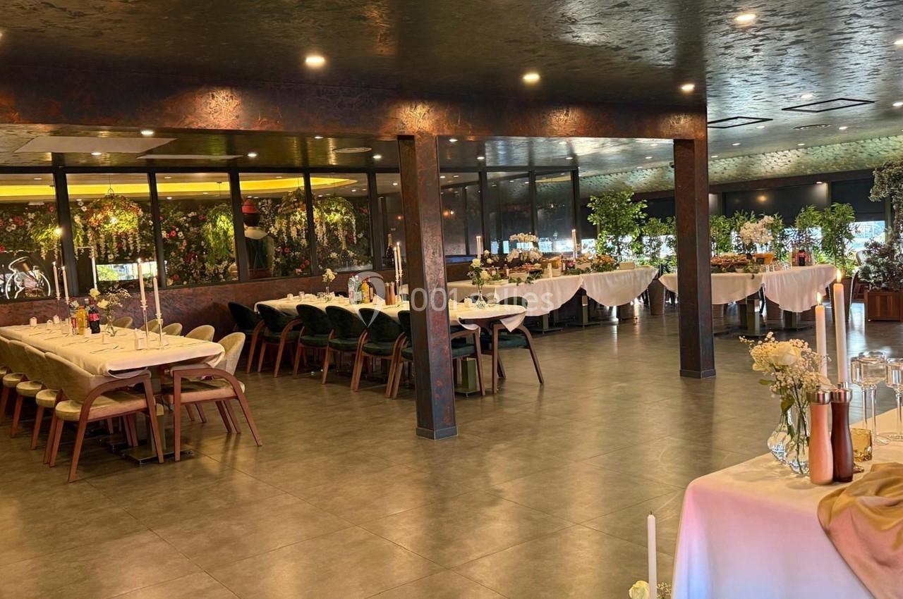 Salle de réception élégante avec tables dressées, chaises en bois et décoration florale, éclairée par des lumières tamisées.