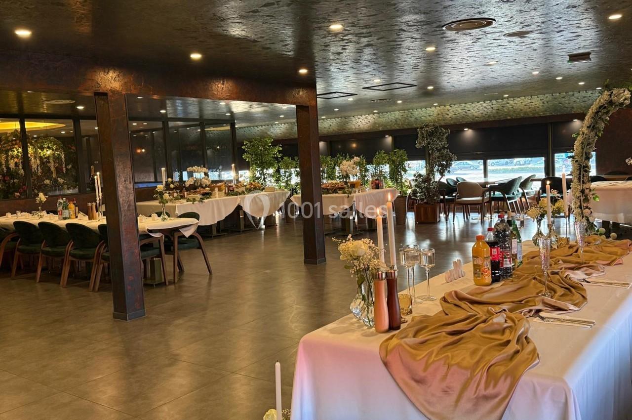 Salle de réception élégante avec tables décorées, nappes dorées et bougies, entourée de grandes baies vitrées.