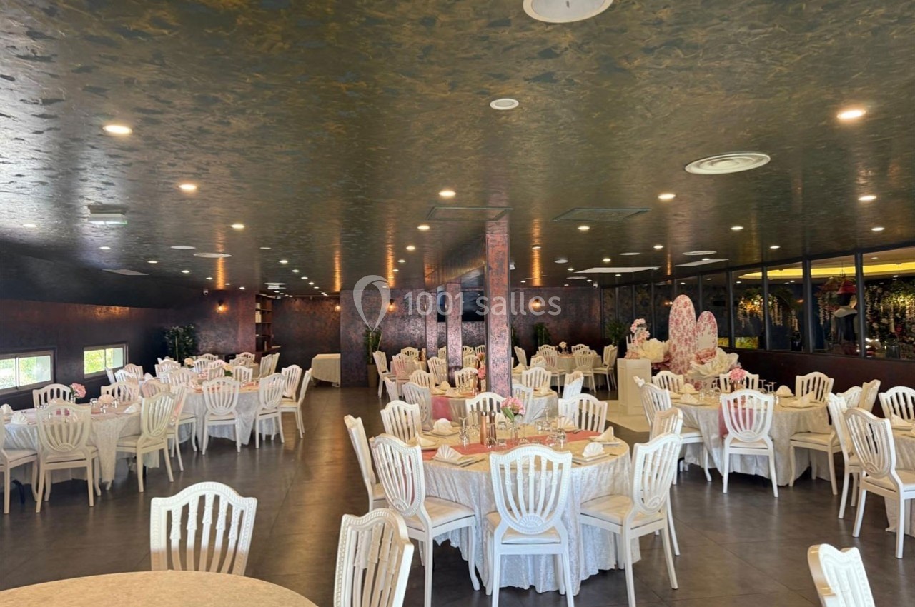 Salle de réception élégante avec tables rondes dressées, chaises blanches et décoration raffinée.