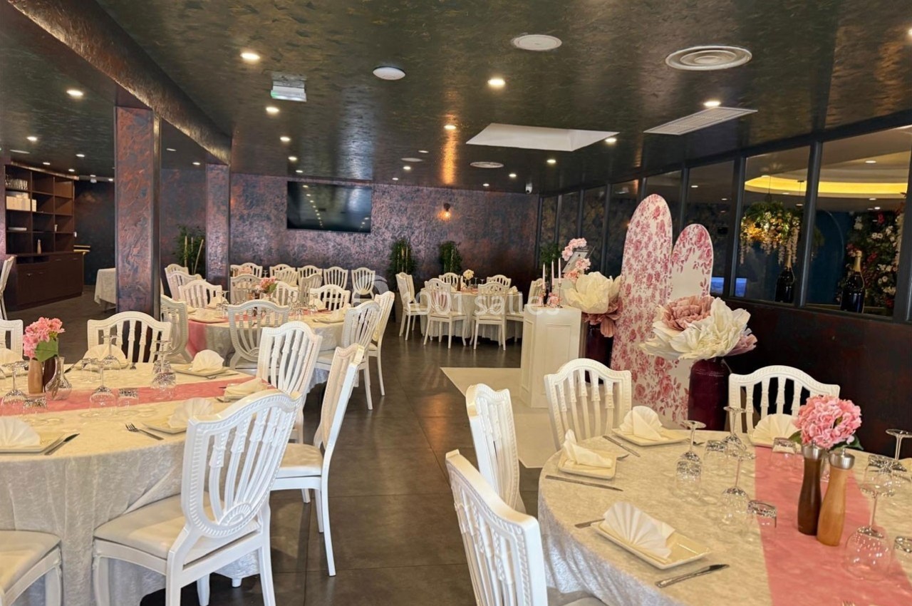 Salle de réception élégante avec tables rondes dressées, nappes beiges, chaises blanches et décorations florales roses.