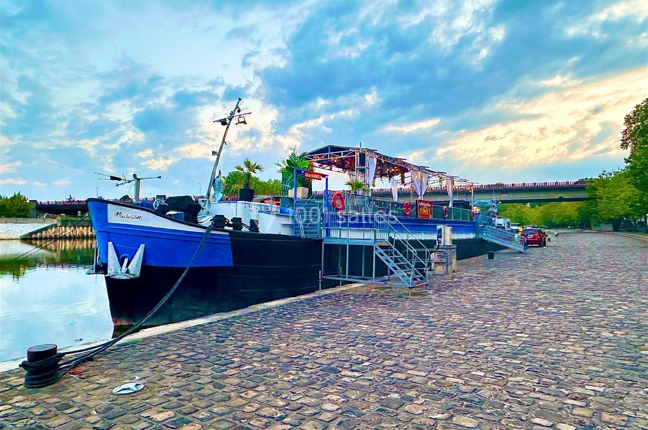 Bateau amarré sur une berge pavée au bord d'une rivière, sous un ciel partiellement nuageux.